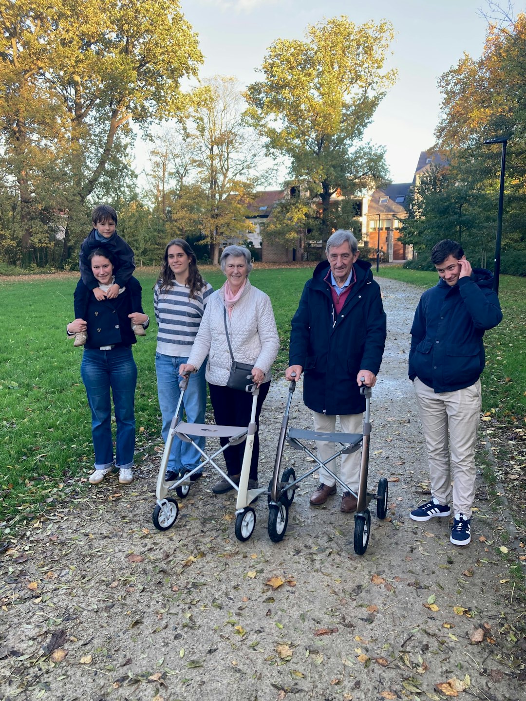 A multi-generational family walks on a park path with two elders using walkers.