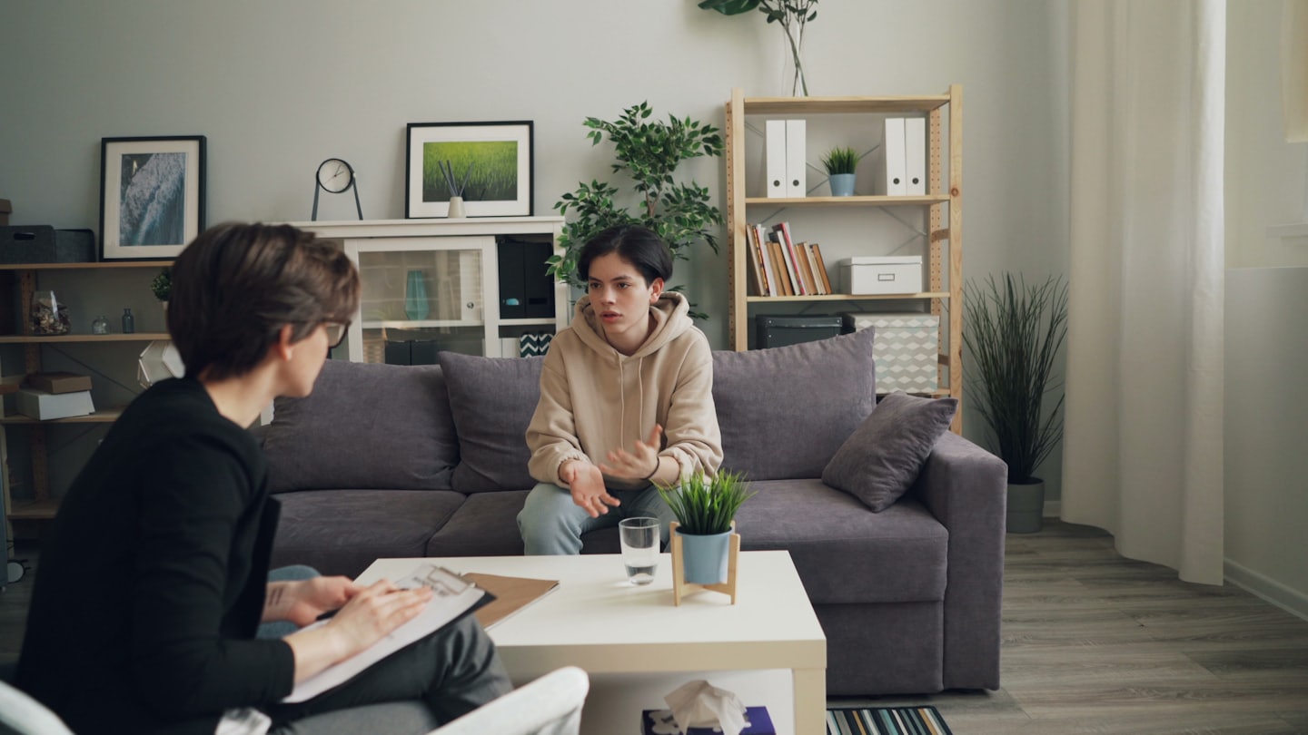 Counselor with clipboard listens to client speaking on sofa.