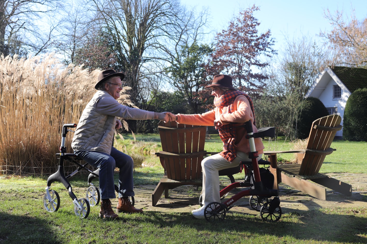 Elderly couple smiling and shaking hands while seated on rollators in a garden.
