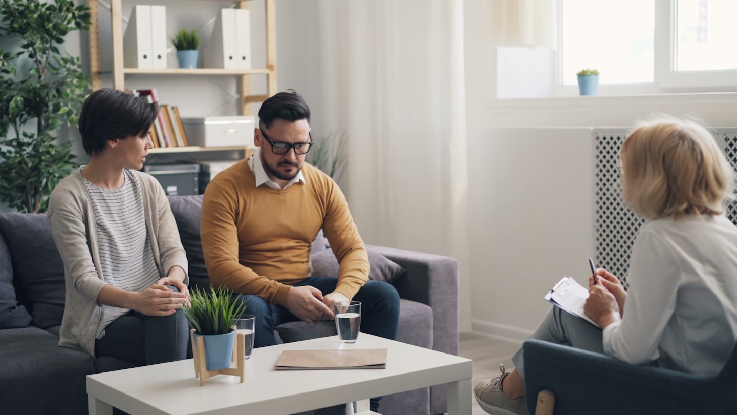 Therapist takes notes while a couple sits on a grey sofa.